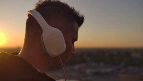 Close-up-of-a-man-in-headphones-listening-to-music-on-the-roof-at-sunset-with-a-view-of-the-city